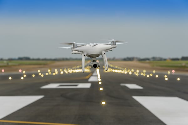 A white drone with a camera hovering above an airport runway.