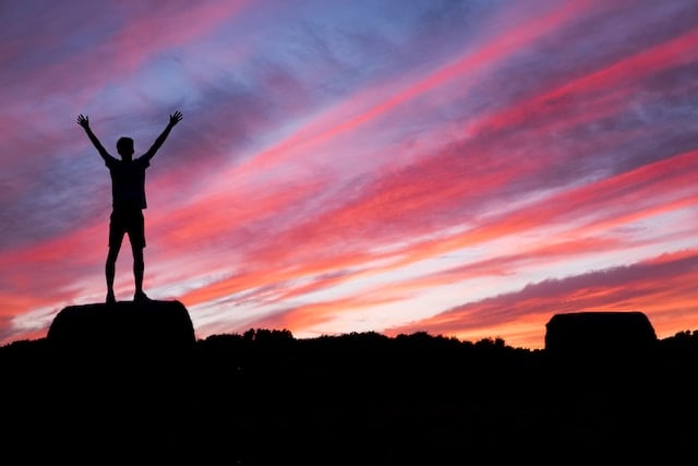 A person standing on top of a rocky surface with a red, blue and orange sky behind them. The person has their hands in the air in a triumphant pose.